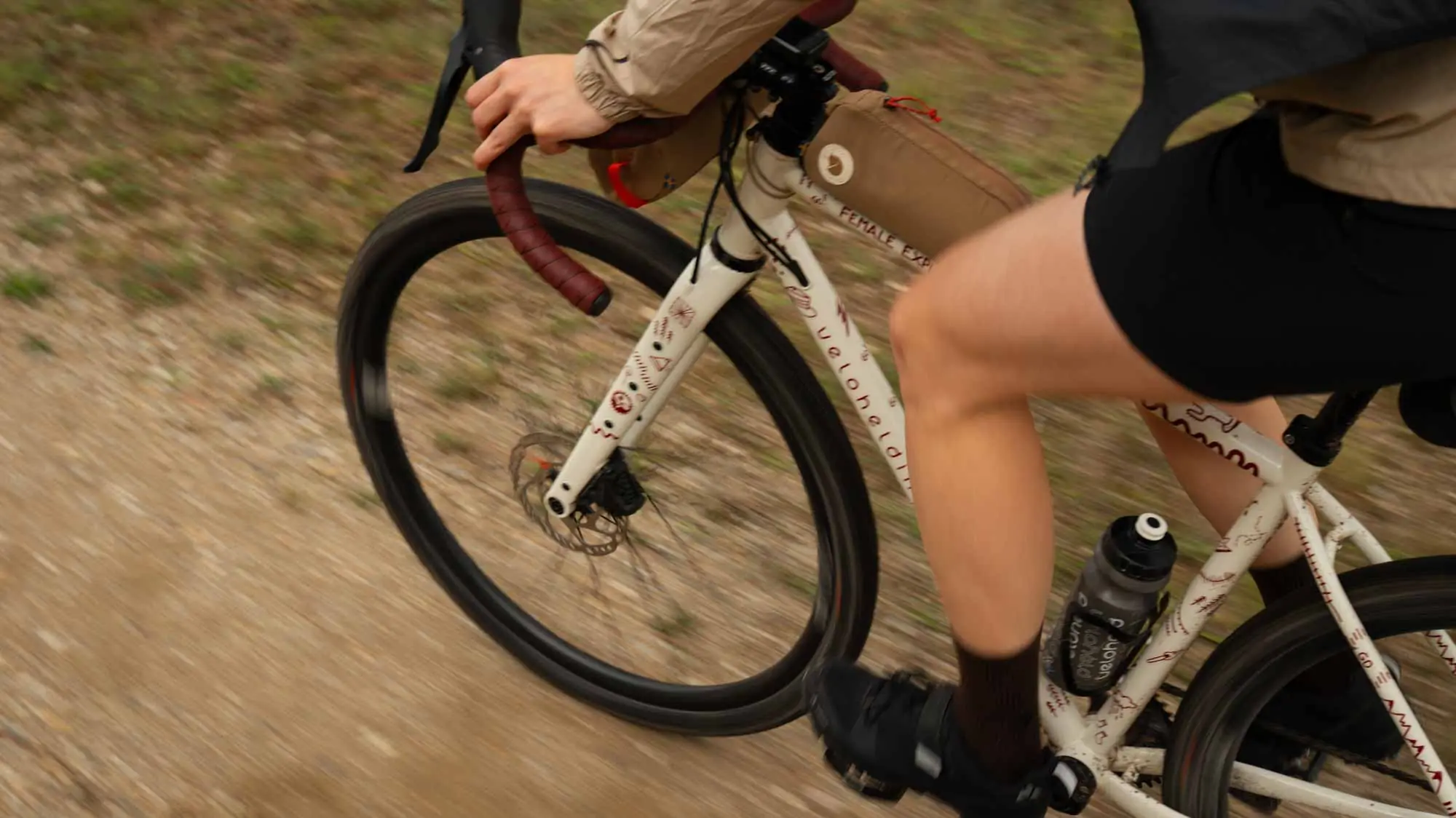 A close-up of a gravel bike being ridden over gravel, a woman is sitting on the gravel bike and you can see her legs and her hands