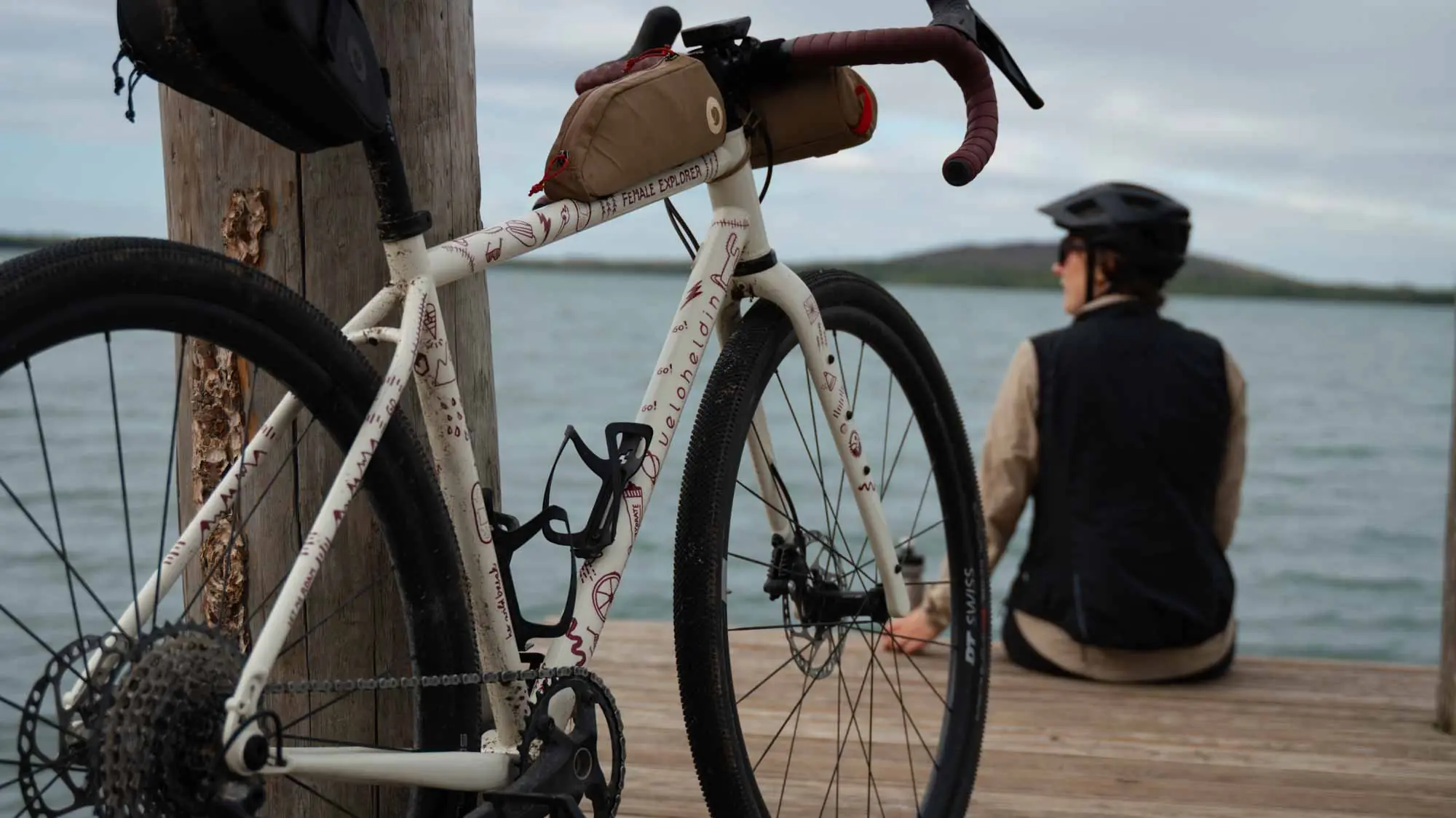 Gravel bike is parked on the jetty, a woman is sitting somewhat away from it at the end of the jetty and looks at the water