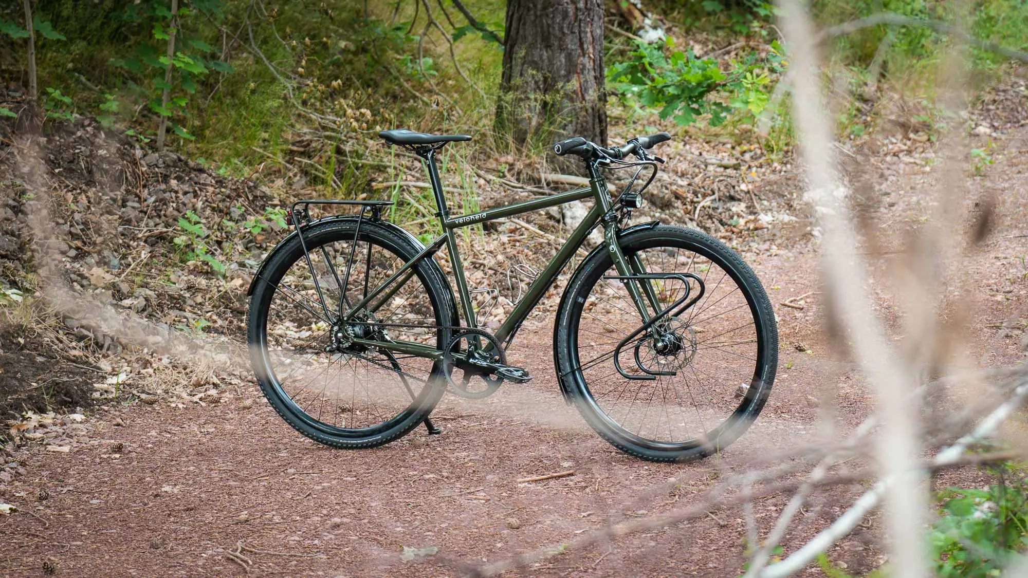 A green veloheld.alleyX with a Rohloff hub gear, Gates belt drive, mudguards and a front luggage rack on a forest path, surrounded by trees and natural scenery.